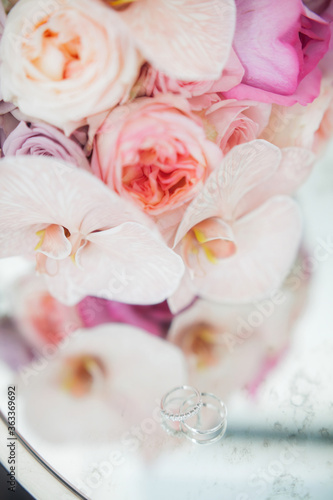 Wedding bouquet of pink and white roses with orchids. Nearby are wedding rings on a mirrored table.