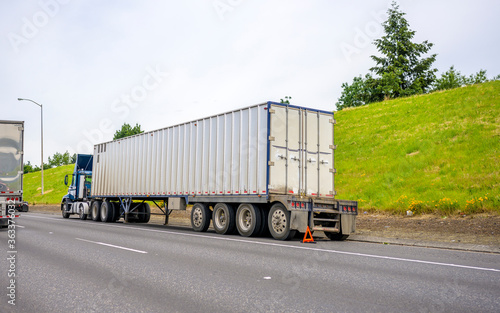 Big rig blue semi truck with loaded container on semi trailer standing broken on the road shoulder set an emergency triangle stop sign behind