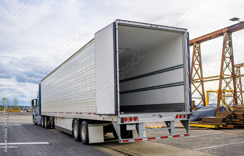 Big rig blue semi truck with open door semi trailer standing at warehouse parking lot at industrial area waiting for the load for the next delivery