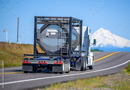 Big rig semi truck transporting flammable chemical cargo in a specialized reinforced tank on open bed semi trailer running on the narrow road with snow mountain view