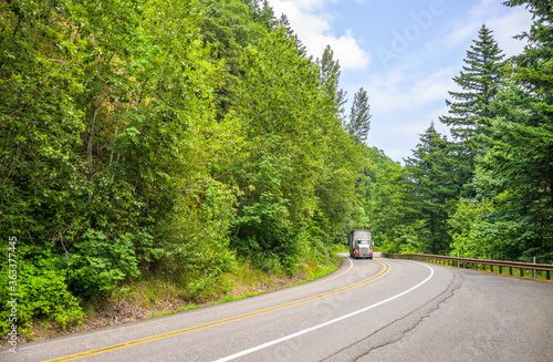 Big rig semi truck with orange fenders transporting cargo in semi trailer running on the winding road through the green forest in Columbia Gorge National scenic area