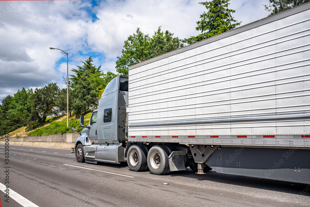 Stylish professional gray big rig semi truck transporting frozen cargo in refrigerator semi trailer running on the straight road at sunny day