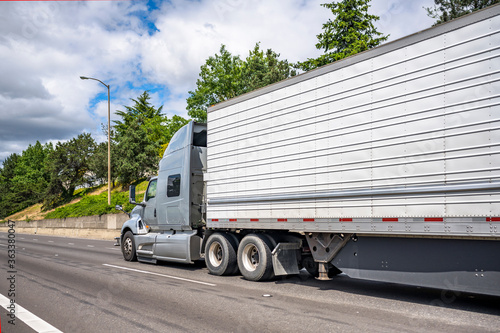 Stylish professional gray big rig semi truck transporting frozen cargo in refrigerator semi trailer running on the straight road at sunny day