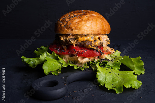 big burger with green lettuce on black background, bun with poppy seeds and sesame seeds, sauce of cheese and vegetables inside,