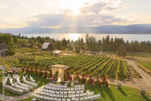 Wedding in the winery. Beautiful romantic set up. Decorated chairs and wedding arbor in front of vineyards by Okanagan lake in Kelowna. The sun shining through clouds during sunset in the background. © Klara