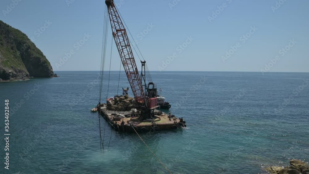 Riomaggiore, Liguria, Italy. June 2020. Outside the marina a floating platform with a dredger is doing maintenance on the artificial rocky barrier. Riomaggiore, Liguria, Italy. June 2020. Outside the marina a floating platform with a dredger is doing maintenance on the artificial rocky barrier.