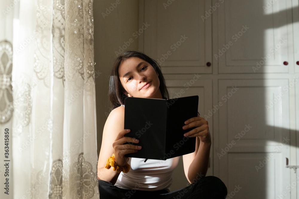 Young woman covering her face with a black book, Hispanic woman in her room smiling looking at the camera, woman enjoying the afternoon with a book Young woman covering her face with a black book, Hispanic woman in her room smiling looking at the camera, woman enjoying the afternoon with a book