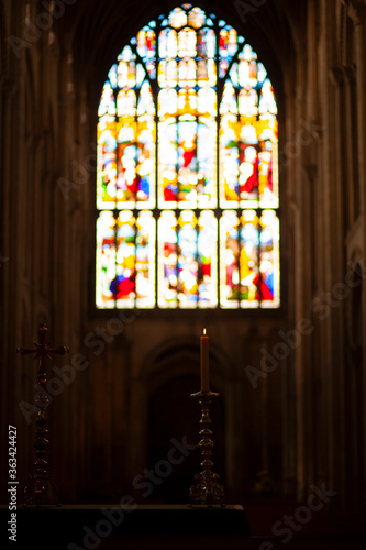 Inside of a historic church in England. Image shows a lit candle and a cross both placed on a table. In the background there is an arched window with vibrant fresco glass paintings. Taken at low light photo