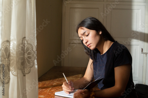 Young Hispanic woman writing in a notebook, serious girl writing in her journal in her room at sunset. concentrated young woman studying at home