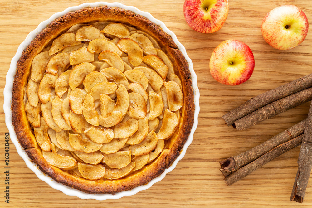 Fototapeta premium Homemade baked French apple tart, an open faced apple pie, in a baking white ceramic dish aside Gala red apples and cinnamon sticks, all on an oak wood background. Flat lay, top view.