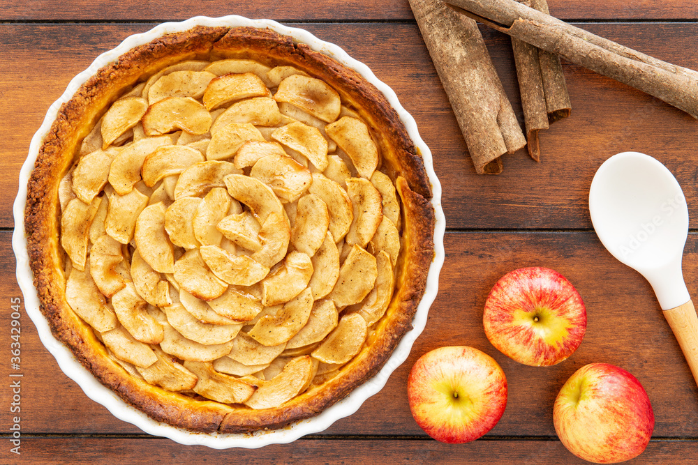 Homemade baked French apple tart, an open faced apple pie, in a baking white ceramic dish aside Gala red apple, cinnamon sticks and white cooking spoon on a vintage wood background. Flat lay, top view
