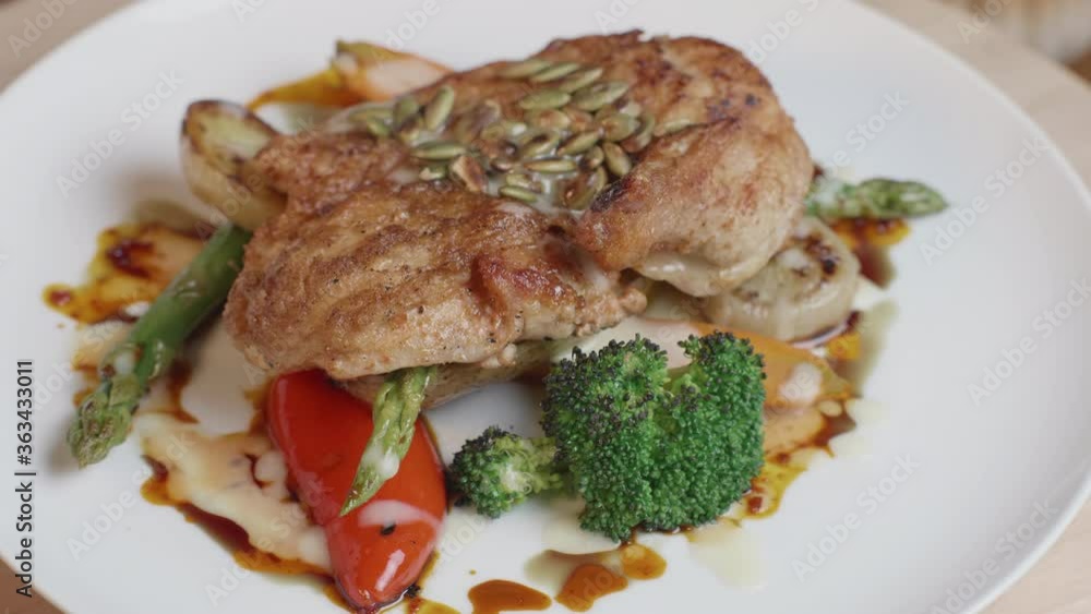 A well presented chicken cutlet, perfectly seared and roasted, topped with pumpkin seeds, and laying on top of colorful vegetable greens. Rotating shot A well presented chicken cutlet, perfectly seared and roasted, topped with pumpkin seeds, and laying on top of colorful vegetable greens. Rotating shot
