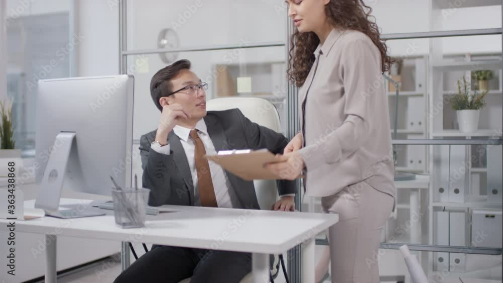 Elegant young woman with long curly hair bringing papers to her cheerful young boss and talking about stats, slow motion shot Elegant young woman with long curly hair bringing papers to her cheerful young boss and talking about stats, slow motion shot