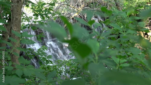 Panning through tree branches and leaves with view of a small man made waterfall cascading down a rocky cliff into the river below. photo