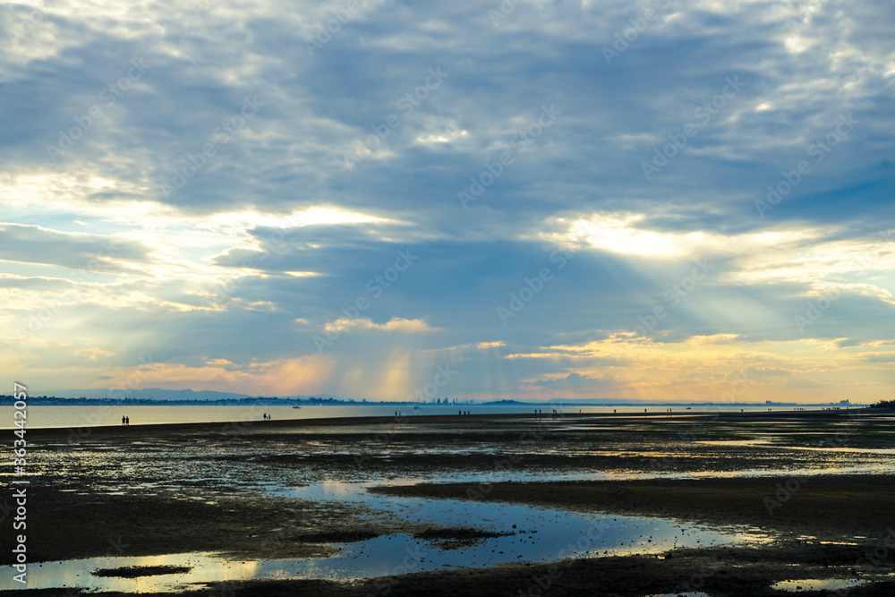 Obraz premium Sunset over Moreton Bay, with light streaming down from behind grey clouds, and people walking to King Island at low tide. Wellington Point.