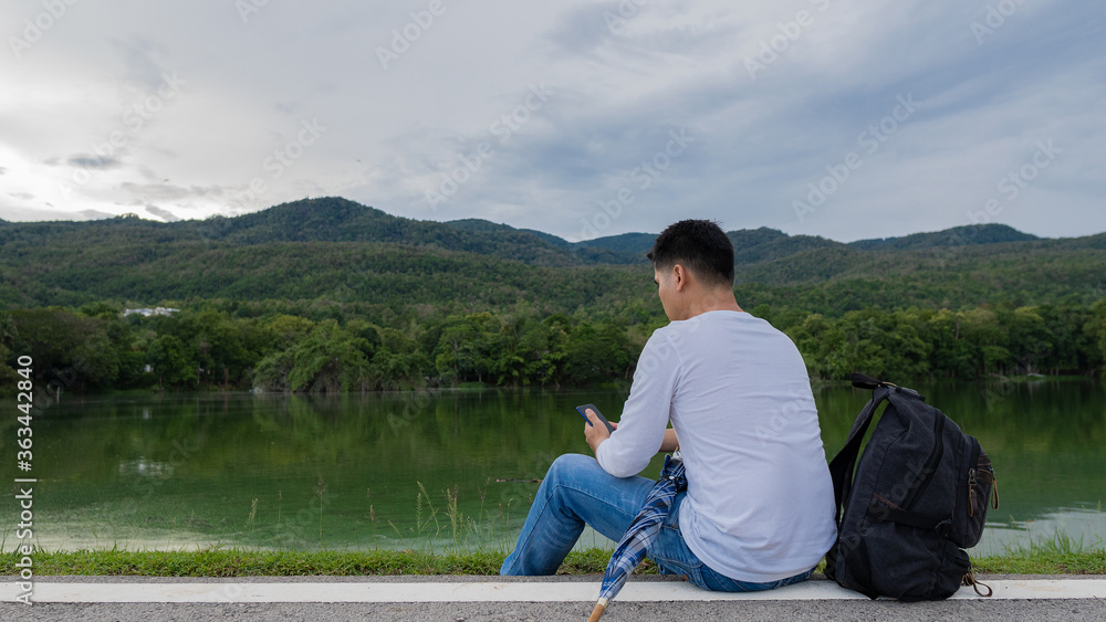 The man sat by the river and played with a mobile phone, with an umbrella and a bag beside him, with a beautiful mountain and sky.