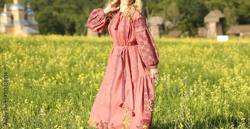 Caucasian girl dressed in a traditional costume with embroidery walks in a field where yellow flowers are on a summer day. Traditions. Summer. Fashion and beauty concept.