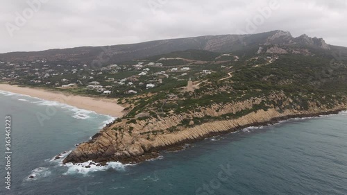 The Camarinal lighthouse on the Atlantic Ocean in Andalusia on a cloudy day. The mountains are forested. In the end you can see the African coast near Gibraltar in the haze.