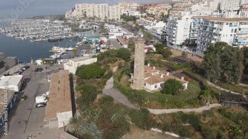 The Punta Doncella lighthouse stands at the port of Estepona. In the background are the boat marina and the houses of the city. The lighthouse is surrounded by trees.