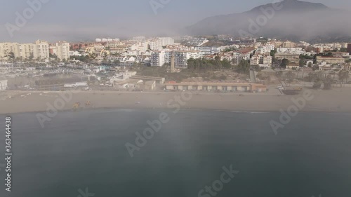 The coast and the beach of the city of Estepona in Andalusia in the morning in the fog. In front of it is the lighthouse Punta Doncella. There is fog and haze over the sea and behind the city.