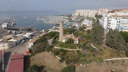 The Punta Doncella lighthouse stands at the port of Estepona. In the background is the boat marina. There is fog and haze over the sea and behind the city.
