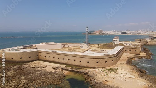 The small fortress island of San Sebastian in the Atlantic Ocean in front of the Spanish port city of Cadiz. A sea side aerial view with a blue sky. The drone flies a curve towards the lighthouse.