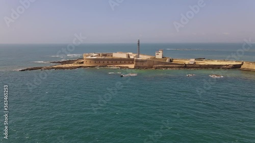 Aerial view of the small fortress island of San Sebastian in the Atlantic. The fortress with the lighthouse lies in front of the Spanish port city of Cadiz. The drone is flying towards the lighthouse.