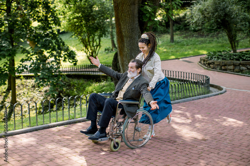 Care helper woman  pretty young hipster granddaughter walking with happy elderly disabled man with arms outstretched  pushing a wheelchair and running in the city park in summer day.