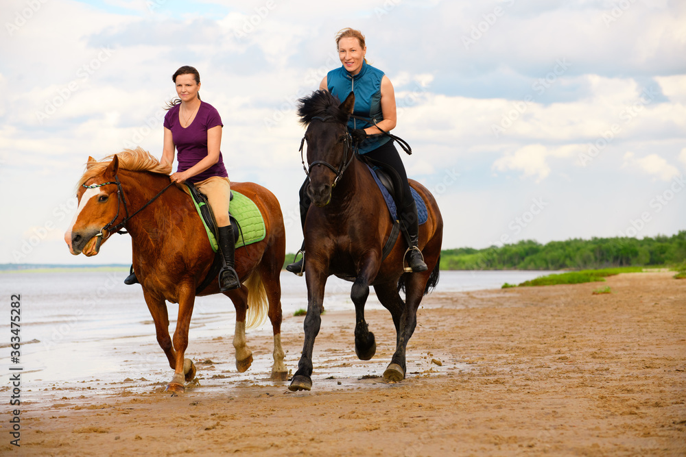 Two happy horsewomen are riding along the beach near the water.