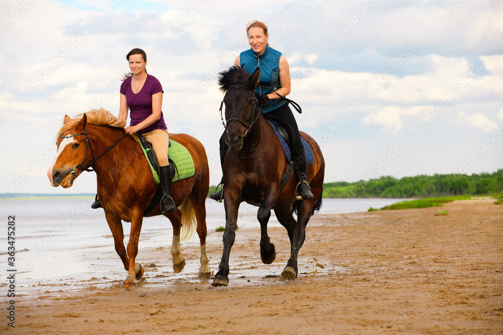 Two happy horsewomen are riding along the beach near the water. Two happy horsewomen are riding along the beach near the water.