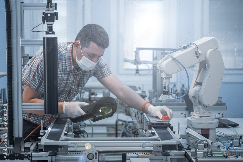 Man with surgical mask and gloves against COVID-19 ( Coronavirus ) programming robot arm with teach pendant. industry 4.0 : smart factory production line control with automation. Man with surgical mask and gloves against COVID-19 ( Coronavirus ) programming robot arm with teach pendant. industry 4.0 : smart factory production line control with automation.
