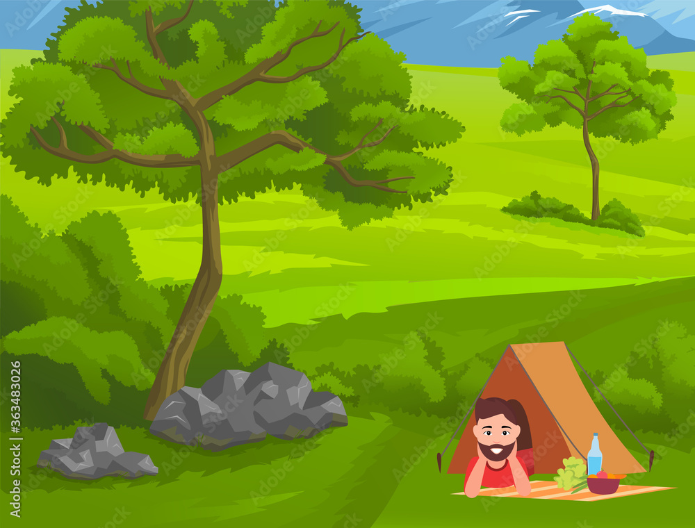 Young guy relaxing lying under tent at green grass at foot of mountains near tree. Happy tourist enjoy nature and recreation. Picnic food such as salad, vegetables, bottle with water at blanket