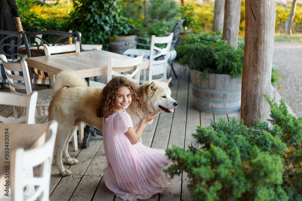 Cute little girl with a big white dog sitting on terrace balcony. A beautiful 7 year old girl in pink dress hugs her favorite dog during a summer walk. Cute little girl with a big white dog sitting on terrace balcony. A beautiful 7 year old girl in pink dress hugs her favorite dog during a summer walk.