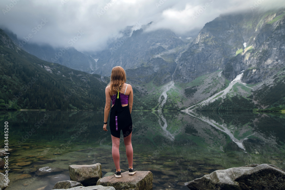 Rear view shot of young fit woman in sportswear looking on the scenic green hills and mountains in dark clouds on the Marine Eye lake, High Tatras, Zakopane, Poland. Foggy day