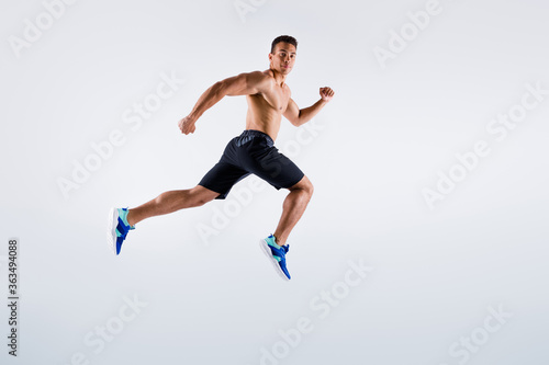 Full length profile photo of handsome sportsman dark skin guy naked chest jumping high up running fast sprint workout exercise wear shorts sneakers isolated white color background