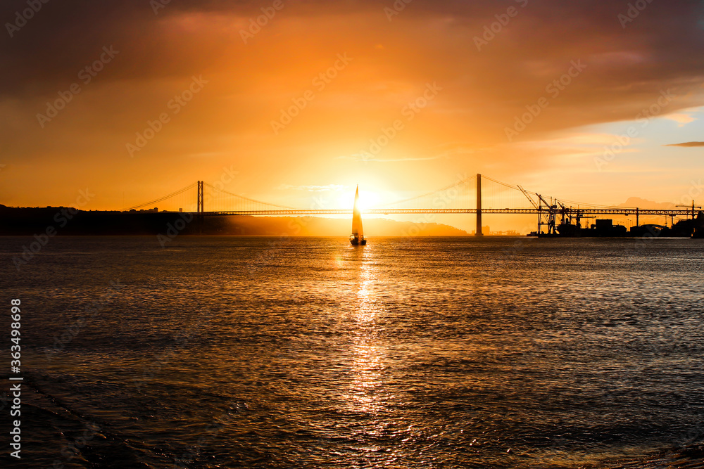 sunset, sailboat stands in front of the sun, reflection on the sea, bridge and buildings behind