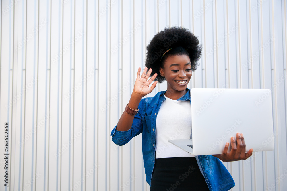 Close-up portrait happy african american young woman with dark curly hair holding in hands laptop and looking at him, making a video call, over grey background in city, empty copy space. Close-up portrait happy african american young woman with dark curly hair holding in hands laptop and looking at him, making a video call, over grey background in city, empty copy space.