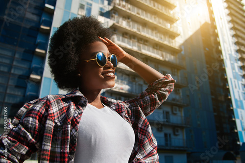 Positive emotions and people concept - young happy black african american woman with curly black hair, wearing sunglasses. Over city background. Lifestyle.
