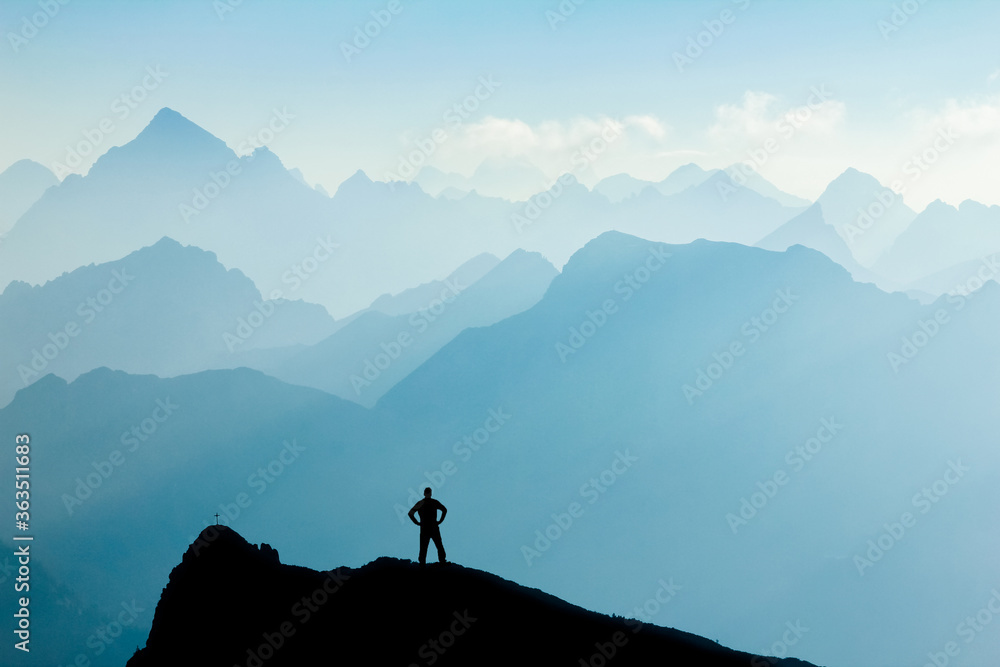 Man reaching summit after climbing and hiking enjoying freedom and looking towards mountains silhouettes panorama during sunrise.