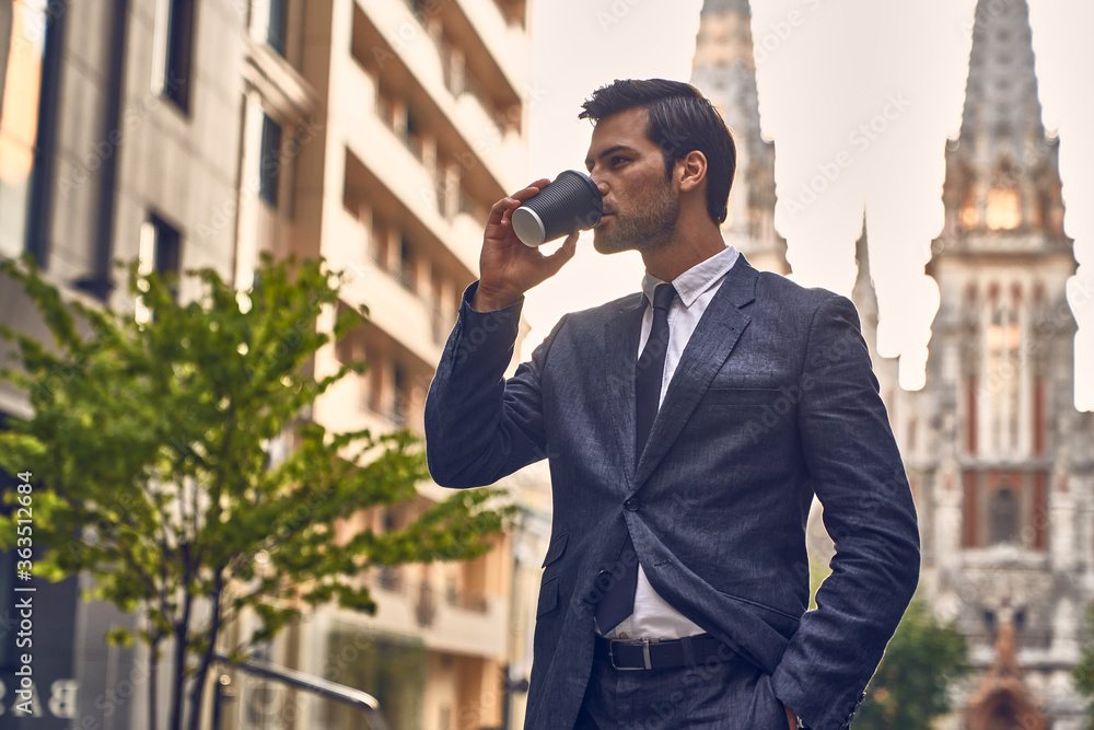 Coffee break. Confident young man in full suit holding coffee cup and looking away while standing outdoors with cityscape in the background. Coffee break. Confident young man in full suit holding coffee cup and looking away while standing outdoors with cityscape in the background.