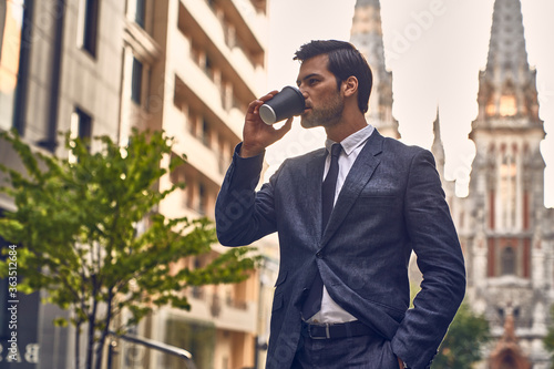 Coffee break. Confident young man in full suit holding coffee cup and looking away while standing outdoors with cityscape in the background. © Roman