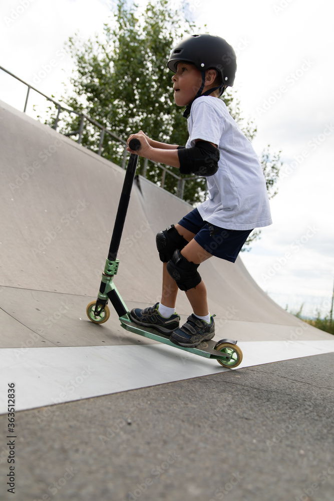 A cute little boy rides a scooter in a skatepark.