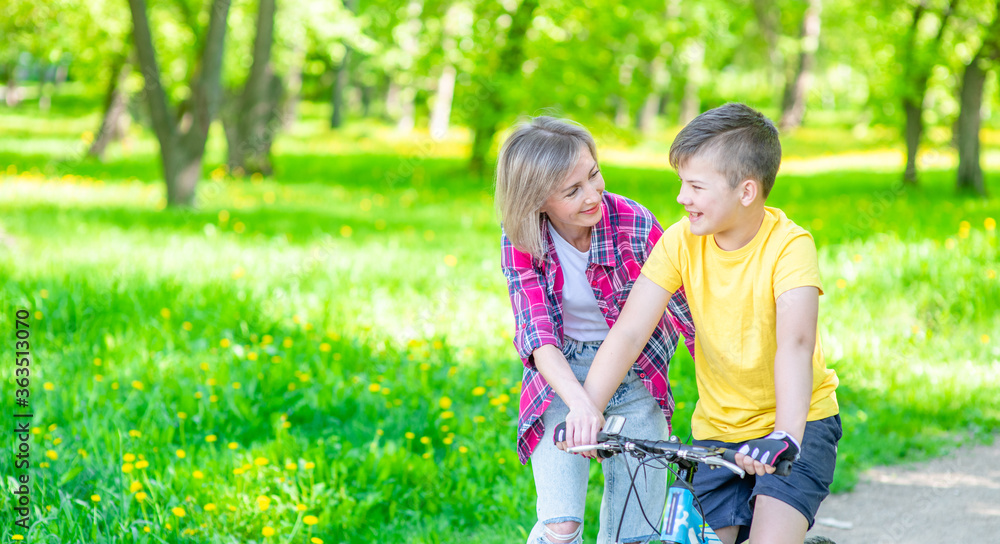 Fototapeta premium Happy family. Mom talks with her young son who ride a bike in summer park. Empty space for text