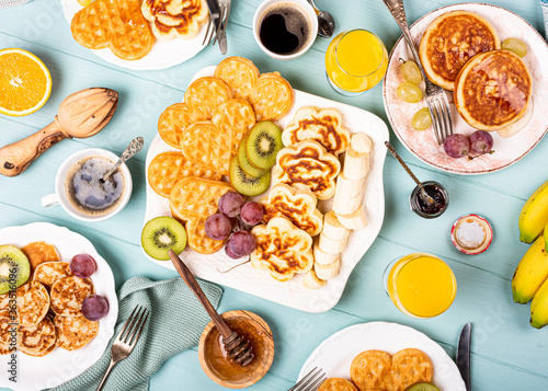 Flat lay with healthy breakfast with fresh hot waffles hearts, pancakes flowers with berry jam and fruits on turquoise background, top view, flat lay. Food concept.