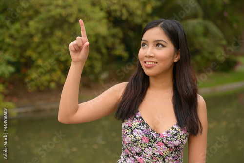 Portrait of happy young beautiful Asian woman pointing finger at the park