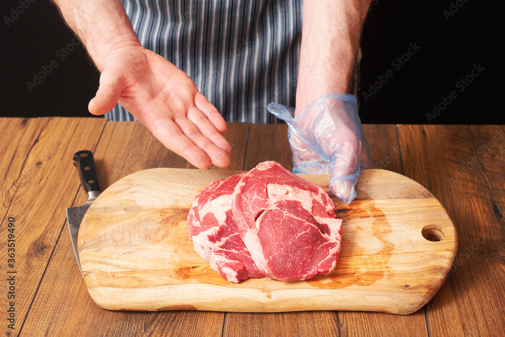 Butcher holding wooden cutting board with two fresh raw rib eye steaks. Right hand pointing to the meat.