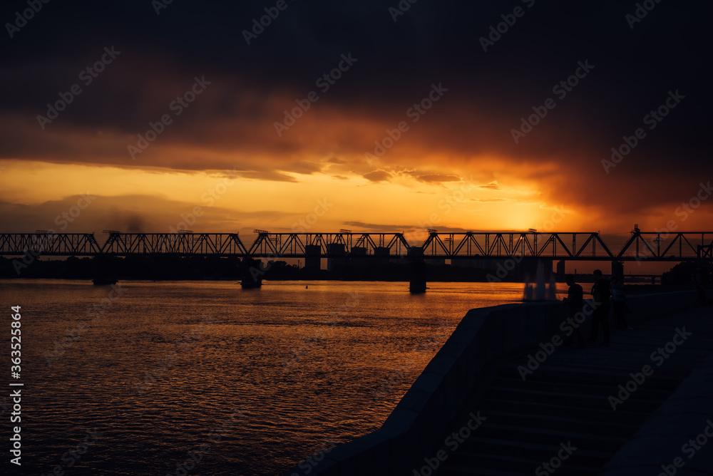 Urban evening landscape. Railway bridge over the river at sunset. Fantastic clouds in the dark sky.