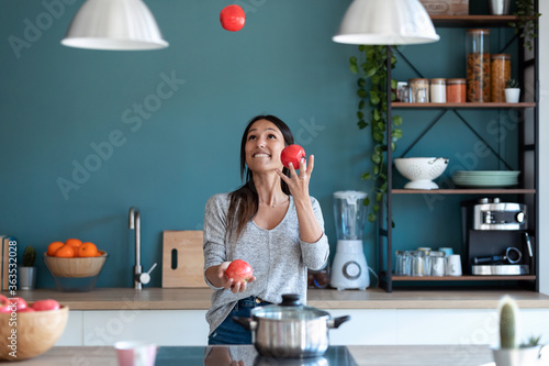 Billede på lærred Funny young woman juggling with three red apples in the kitchen at home