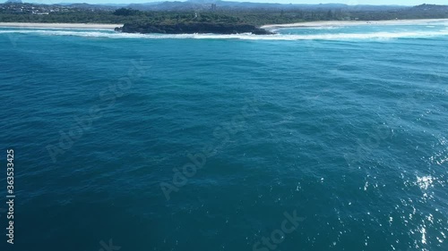 Aerial approach over ocean to small lighthouse on headland with volcanic rock cliffs.