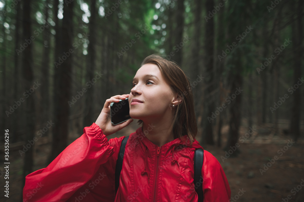 Cheerful attractive tourist woman is talking on the phone in the mountain fir forest. Positive hiker girl in a red jacket is speaking on the smartphone in the woods. Closeup photo. Cheerful attractive tourist woman is talking on the phone in the mountain fir forest. Positive hiker girl in a red jacket is speaking on the smartphone in the woods. Closeup photo.